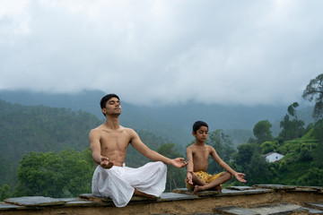 A young handsome boy doing yoga with a kid on the roof of a house situated in the middle of mountain range. Yoga and fitness.