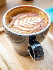 Hot milk coffe, latte, with milk foam art top, in black ceramic cup, served on wooden plate in morning cafe