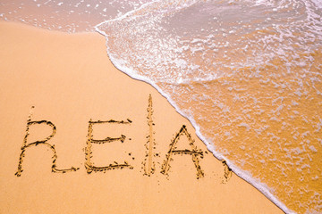 Word Relax hand written in the sand with a sea wave. Close up sand texture on beach in summer.