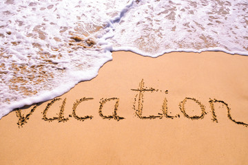 Word Vacation hand written in the sand with a sea wave. Close up sand texture on beach in summer.