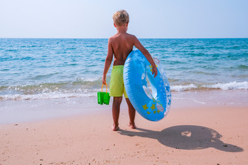 Happy little boy standing near the sea with a blue and yellow pool float in his hands. Copy space banner.