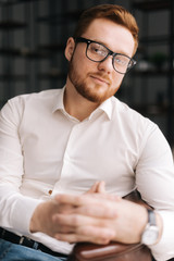 Confident young business man wearing stylish glasses in trendy clothing looking at camera while sitting indoors on chair in modern office. Portrait of bearded handsome gentleman wearing white shirt.