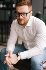 Smiling young business man wearing stylish glasses in trendy clothing looking at camera while sitting indoors on chair in modern office. Portrait of bearded handsome gentleman wearing white shirt.