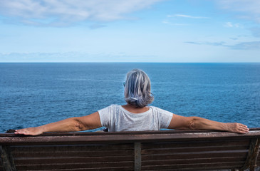 Fototapeta premium Back view of senior woman sitting on a wooden bench enjoying relax and nature. Blue sea and sky on background - positive moment during happy retirement