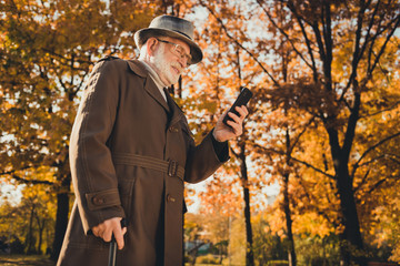 Low angle view profile photo of retired cheerful white hair grandpa walk desert park stick chatting telephone family good mood look forward meet them wear specs coat cap autumn colors outdoors