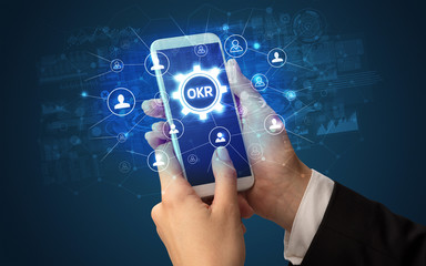 Female hand holding smartphone with OKR abbreviation, modern technology concept
