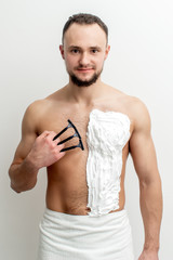 Young caucasian man with beard holds razor shaves his chest with white shaving foam on white background. Man shaving his torso