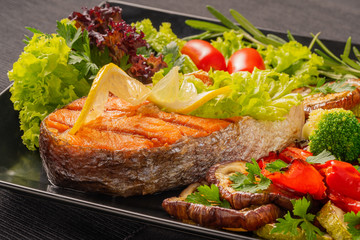 Serve food in the restaurant. Food poster - large portion of red fish, trout, salmon steak with grilled vegetables, salad, broccoli and tomatoes, decorate with lemon and rosemary.