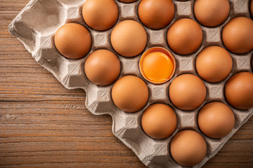 Many fresh raw brown chicken eggs in carton. Beautyful brown chicken egg is half broken with yolk among other eggs, top view on wooden background. Top view.