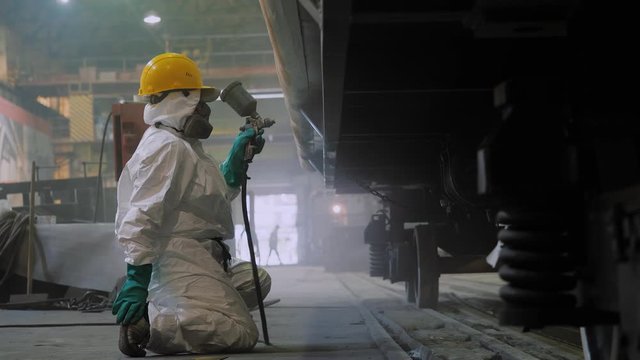 Factory worker in protective suit paints surface of train wagon with spray gun