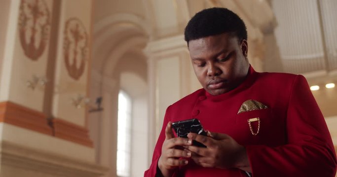 Portrait of african american man in red suit using his smartphone. Young male person looking and touching phone screen while searching gospel song words. Indoors