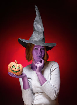 Halloween. A young woman with purple skin in a black witch hat is holding a pumpkin with a zombie face. Dark red background. Vertical