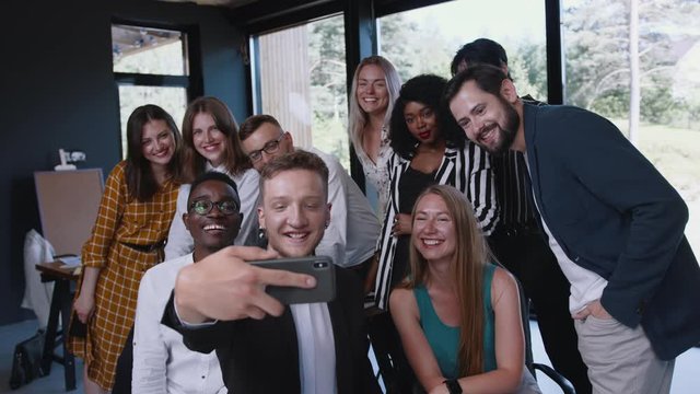 Team of young smiling diverse business people waving together to smartphone selfie camera at modern office workplace.