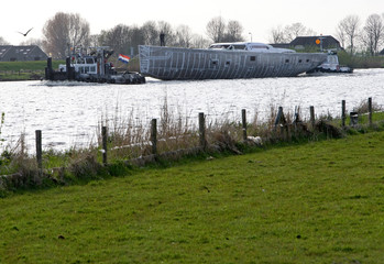Obraz na płótnie Canvas Super sailing yacht aluminium casco transport on a pontoon. Ship building industry. Tugboat. Canal. Genemuiden Netherlands.