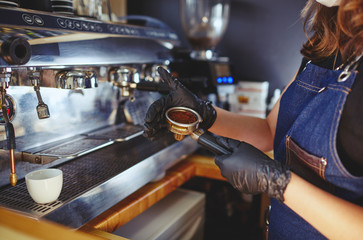 Barista wearing medical latex black gloves,preparing coffee drink with espresso machine in coffee shop cafe.Woman presses ground coffee using tamper.Blurred image, selective focus