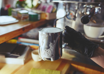 Barista wearing medical latex black gloves, preparing coffee drink with espresso machine in shop cafe. Woman hold metal milk pitcher in hand. Blurred image, selective focus