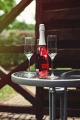 Two flute glasses and an pink champagne bottle with no label on a glass table in a back yard.