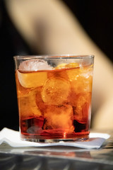 Close up of a glass of Negroni cocktail with gin and vermouth in the outdoor tables of a famous cocktail bar. Ice cuber and orange spirit in the tumbler glass. Adult beverages.