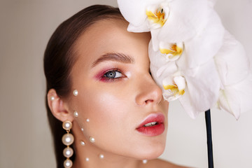 Tender woman with stylish makeup covering eye with orchid flower while looking at camera on gray background 