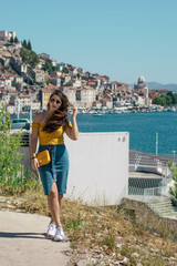 Attractive brunette wearing a blue skirt and an orange top posing outside near the sea on a warm summer day. Long hair flowing in the wind. Outline of Sibenik town in the distance. Influencer travels