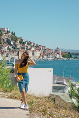 Fototapeta na wymiar Attractive brunette wearing a blue skirt and an orange top posing outside near the sea on a warm summer day. Long hair flowing in the wind. Outline of Sibenik town in the distance. Influencer travels