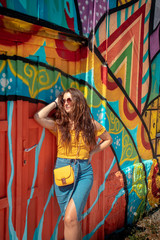 Attractive brunette girl wearing a blue skirt and an orange top posing on a summer day with a colorful wall in the background. Yellow purse, stylish influencer travelling and modelling