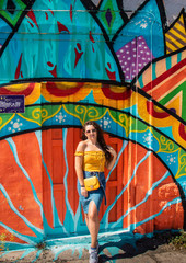 Attractive brunette girl wearing a blue skirt and an orange top posing on a summer day with a colorful wall in the background. Yellow purse, stylish influencer travelling and modelling
