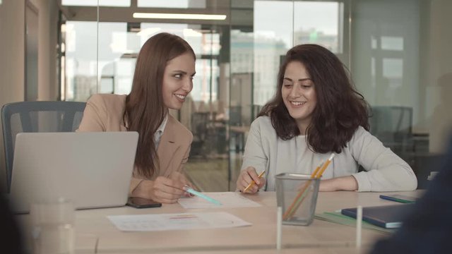 Beck view of two young mixed-race and Caucasian businesswomen sitting at desk in office and discussing document lying on desk with unrecognizable interlocutor sitting in front of them