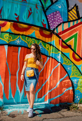 Attractive brunette girl wearing a blue skirt and an orange top posing on a summer day with a colorful wall in the background. Yellow purse, stylish influencer travelling and modelling