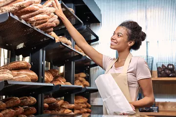 Tuinposter Small Business. Young woman in apron at bakery shop taking baguette from shelf putting into paper bag smiling happy © Viktoriia