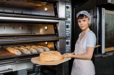 Young caucasian woman baker is holding a wood peel with fresh bread near an oven at baking...