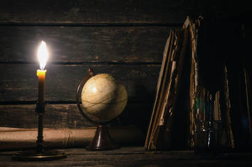 Quill pen in a inkpot, globe and stack of books in the light of burning candle on the writer desk background. Education concept or traveler diary.
