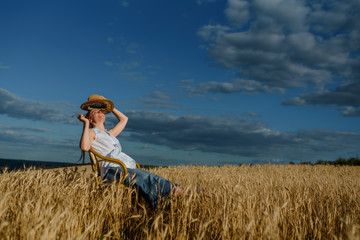 Naklejka na ściany i meble Young woman in casual outfit and a hat is sitting on a vintage chair in the wheat field.
