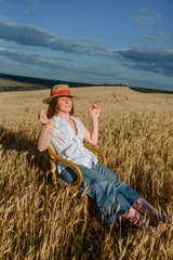 Young woman in casual outfit and a hat is sitting on a vintage chair in the wheat field.