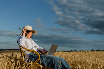 Young woman freelancer is working on a computer outdoors in the wheat field. She is sitting on a vintage chair and enjoying the ability to work from anywhere
