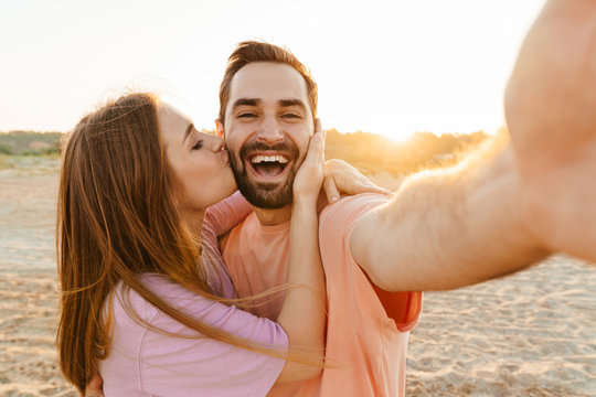 Image of young couple kissing and taking selfie photo by seaside