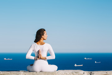 Yoga on high altitude with sea with ships on background, woman seated in yoga pose on amazing sea background, woman meditating yoga enjoying sunny evening, woman makes yoga meditation on mountain hill