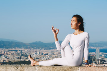 Fototapeta na wymiar Yoga on high altitude with big city on background, young woman seated in yoga pose on amazing city background, woman meditating yoga and enjoying sunny evening, woman makes yoga on mountain hill