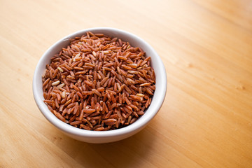 Closeup brown rice or red rice with wooden spoon and white ceramic bowl on wood table top. Healthy organic food concept.