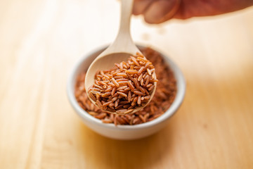 Close up brown rice or red rice with wooden spoon and white ceramic bowl on wood table top. Healthy organic food concept.