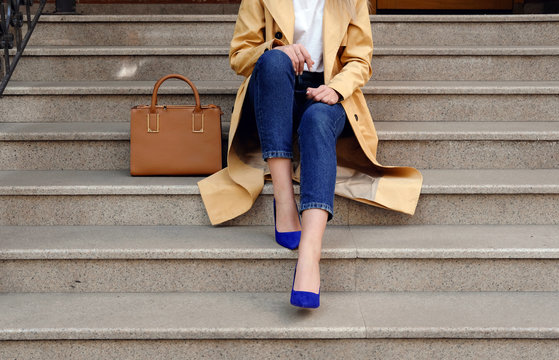 Street fashion outfit, model in blue jeans coat and brown handbag in high heel shoes sitting on the beige stair, outdoor