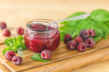 Fresh homemade raspberry jam in glass jar on a wooden background. Several fresh berries and mint are near it.
