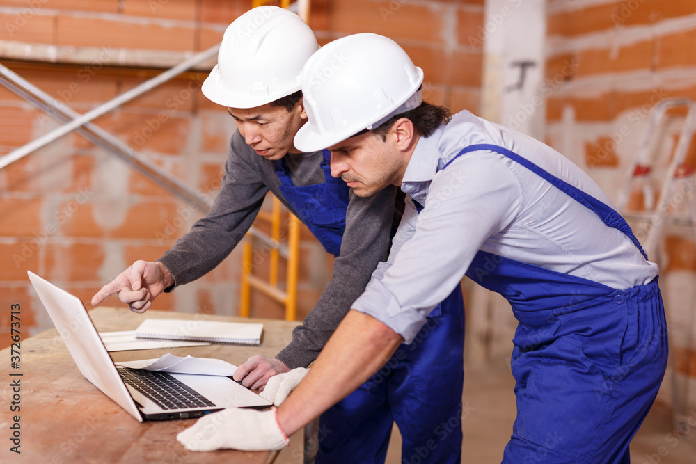 Wall mural portrait of two builders reading plan of brick building using laptop