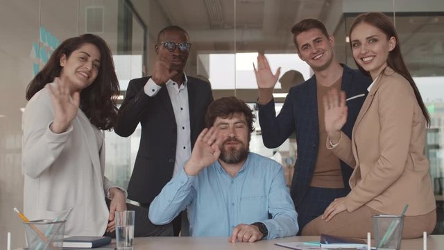 Lockdown of five multiethnic coworkers are gathered around desk in the office, looking at camera and waving their hands