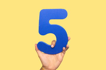 Concept of be the fifth one, fifth birthday, fifth place, losing. Hand holding a number five made of plastic. Blue subject with yellow background