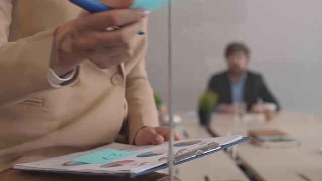 Close up of unrecognizable businesswoman sitting near glass wall in the office and sticking blue stickers on clipboard with chart while man working on background