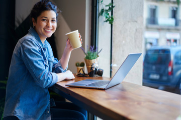 Successful businesswoman looking at camera while holding coffee in hand and working at laptop device indoors.Cheerful web designer enjoying drink during break sitting at netbook computer in cafe