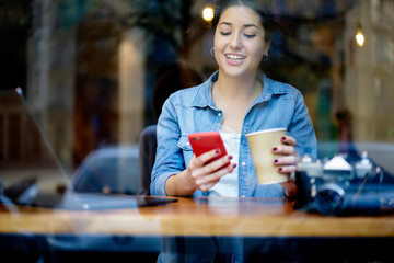 Cheerful hipster girl reading funny sms message on red smartphone sitting in coffee shop with tasty beverage in hand.Positive student chatting on telephone enjoying drink in cafe during leisure time
