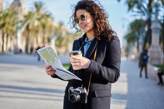 Smiling stylish hipster girl in trendy sunglasses searching right destination on map standing with tasty coffee in hand outdoors.Positive traveller with camera looking for interesting places