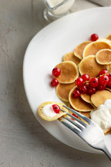 Tiny pancake cereal on white plate served with red currant berries and ingredients on grey cement table. Trendy breakfast, small round baked crepe batter drops. Homemade food. Vertical
